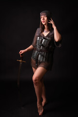 Fototapeta premium Full leg portrait of a beautiful young woman in oriental chainmail armor, holding a saber, posing isolated against a dark background. Concept art of an oriental warrior.
