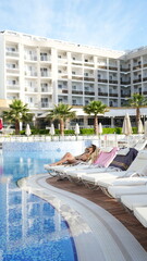 Fototapeta premium Luxury Resort Bliss: Two women relax on lounge chairs by a shimmering blue pool at a modern resort, with a grand white building and palm trees in the background.