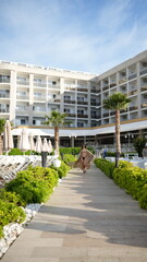 Fototapeta premium Tropical Resort Escape: A woman in a flowing cover-up walks down a scenic path towards a luxurious beachfront resort, palm trees swaying gently under a clear blue sky.