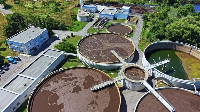 A wastewater treatment plant located by a river features several large circular tanks used for processing water. Surrounding buildings support waste management and treatment tasks during daytime.