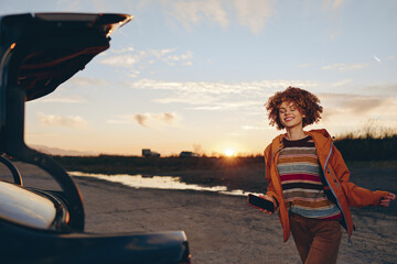 Fototapeta premium Smiling woman with curly hair by a car on a seaside road at sunset, travel lifestyle, warm sky, casual outfit, outdoor moment, soft natural light