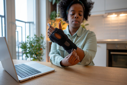 Focused woman adjusting orthopedic wrist brace while sitting at table with laptop. Concept of rehabilitation, injury recovery, carpal tunnel syndrome, chronic pain management and healthcare support.