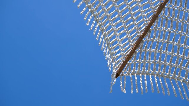 Close-up footage of a white fringed parasol swaying in the wind against a cloudless sky, exotic paradise destination vacation