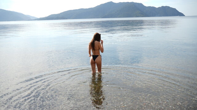 A Woman in Bikini Enters the Ocean: A woman in a bikini stands in the shallow water of a calm ocean, with a tranquil mountain landscape in the background.