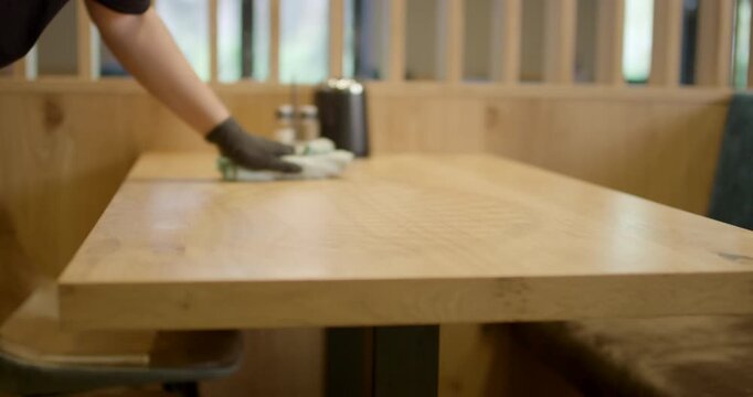 Worker in black safety glove wiping wooden restaurant dining table surface with microfiber rag cloth