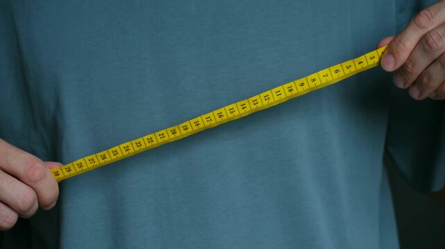 Close up male hands unrolling yellow measuring tape in front of his torso, concept for body measurement, tailoring, weight loss diet tracking in centimeters. Male hands unrolling yellow measuring tape