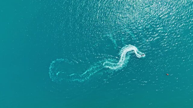 A boat moves through clear blue water, creating waves, while another small object is visible in the distance. The scene is bright and captures a lively summer day on the water.