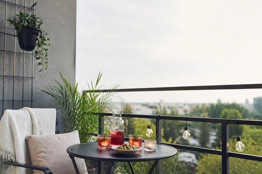 A small outdoor table with drinks and snacks is set on a balcony overlooking a city landscape under an overcast sky.