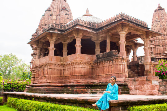 Young woman in a blue dress sitting peacefully in front of the magnificent red sandstone temple at Mandore Garden, Jodhpur, Rajasthan, India.