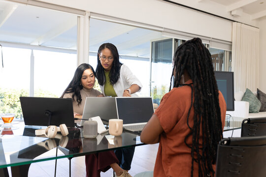 Diverse female colleagues collaborating around glass-topped table in home office using laptops