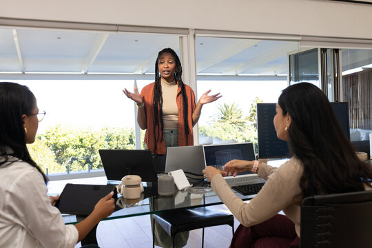Diverse female team meeting at glass table, lead in rust cardigan gesturing with laptops tablet