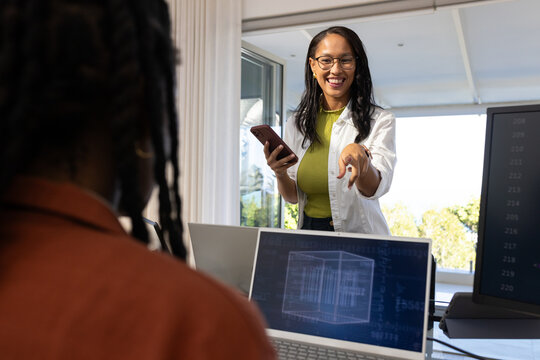 African American women at office in white top blazer pointing, holding phone near laptop screen