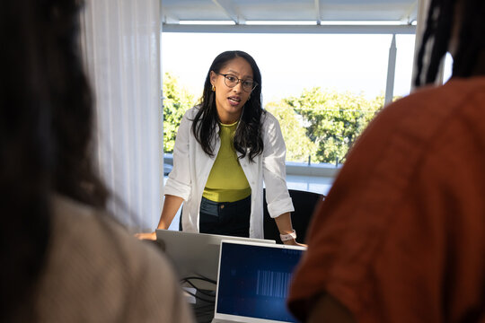 Diverse female coworkers in office, presenter leaning, addressing team, laptops showing barcode