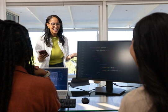 Diverse female team in office listening, leader in blazer holding phone pointing at code monitor