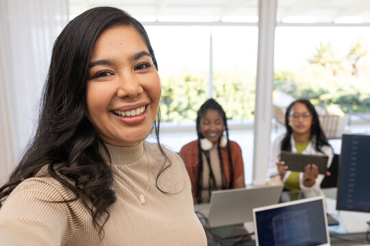 Diverse female coworkers smiling, collaborating in office with laptops, tablet, pendant