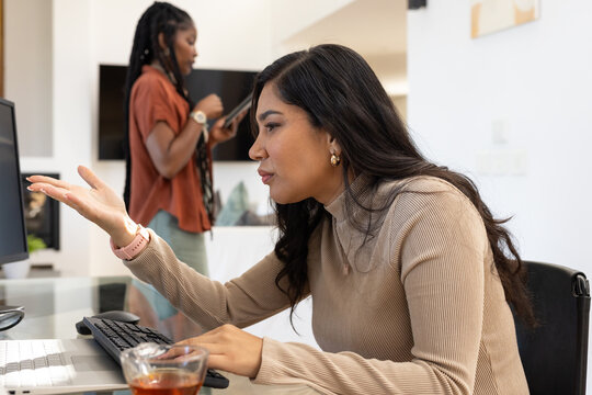 Two female coworkers in office, beige-clad woman typing at keys near glass cup, peer holding tablet