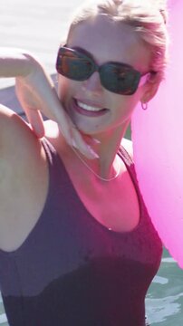 Vertical video: Holding bobbing pink ball, woman posing with waves in water by dock, dark swimsuit