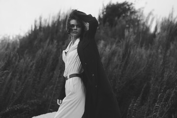 Fototapeta premium Monochrome fashion shot of a woman in a white dress and long coat standing in tall grass, dramatic pose, wind swept hair, outdoor moody portrait with strong contrast