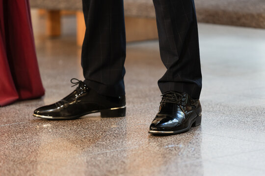 Shiny black dress shoes paired with dark suit trousers on a polished floor, suggesting a formal event, wedding or evening occasion. Elegant footwear detail conveying style, refinement and celebration