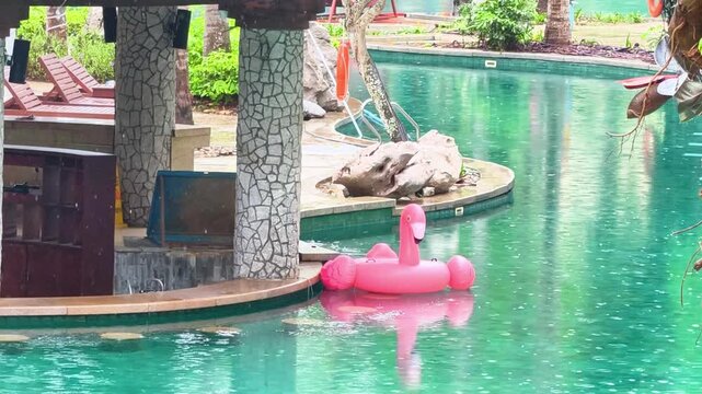 Inflatable pink flamingo floating alone in empty hotel pool during rainfall, tropical resort with palm trees, rainy season vacation, solitude and abandoned leisure concept