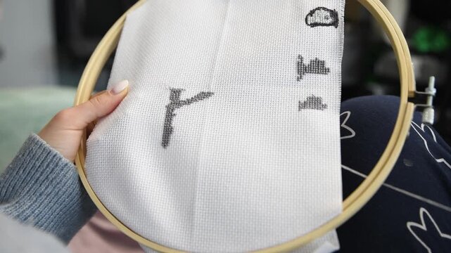 Detailed hands stitching gray cross stitch pattern on white fabric in wooden embroidery hoop indoors