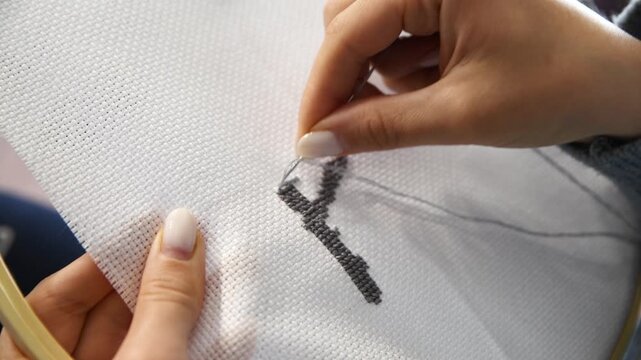 Hands stitching black cross stitch pattern on white fabric with embroidery hoop close up