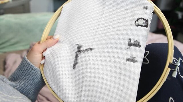 Hand stitching black cross stitch pattern on white fabric with wooden hoop indoors cozy lighting