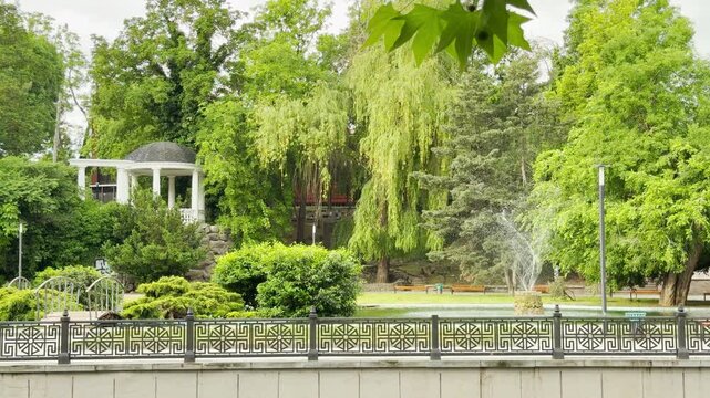 A water fountain sprays water inside a park pond. A white classic rotunda stands on a hill surrounded by lush green trees. A decorative fence lines the foreground.