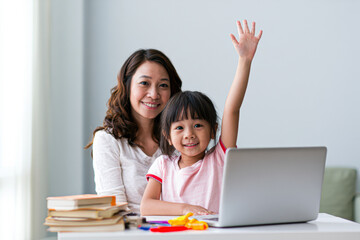 Obraz premium A smiling woman and child sit together at a table with a laptop, books, and stationery, while the child raises their hand enthusiastically.