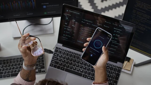High angle cropped shot of hands of unrecognizable programmer holding smartphone with voice assistant app at desk with laptop displaying data. For tech startups promoting voice recognition software