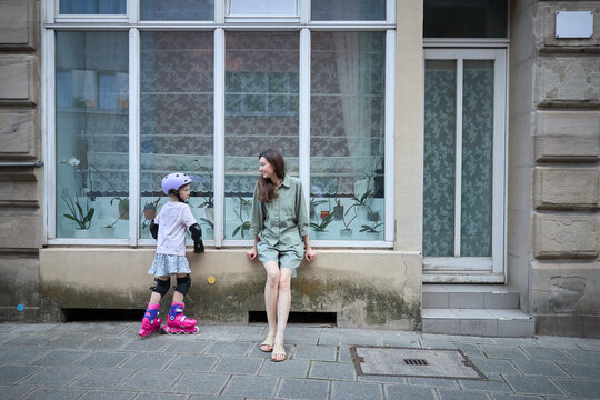 Mother watches daughter in roller skates and protective gear on sidewalk