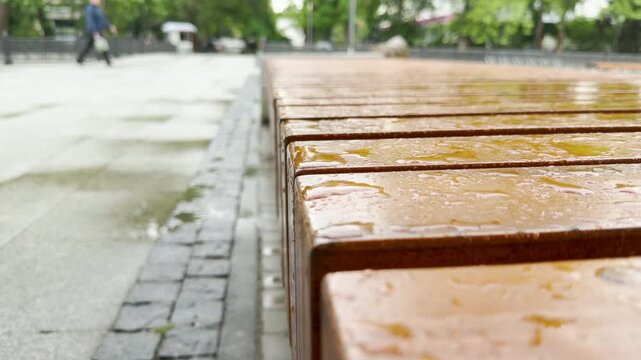 A varnished wooden bench with raindrops sits along a wet stone pavement. A passerby walks in the blurry background of the public square.