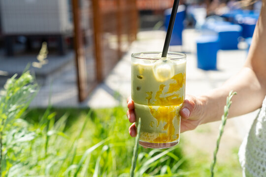 Cropped horizontal shot of a woman holding an iced mango matcha latte drink at a summer cafe terrace