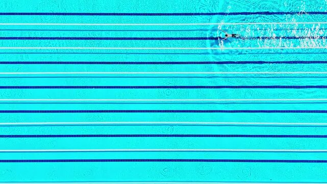 A person swims in a large swimming pool that has blue lane markings on the surface. The water is clear and reflects light. It is bright outside, and the scene shows activity.