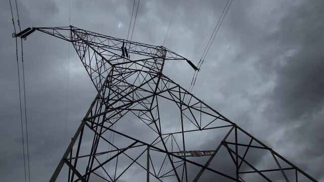 A dramatic, low-angle shot of a power pylon towering against dark overcast clouds. The moody lighting evokes an ominous energy crisis, framing the stark industrial latticework as symbol of grid strain