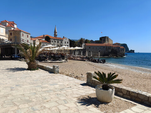 Budva Old Town promenade runs beside sandy beach with seaside cafes and parasols, historic stone buildings and church steeple framing clear Adriatic waters under bright summer sky