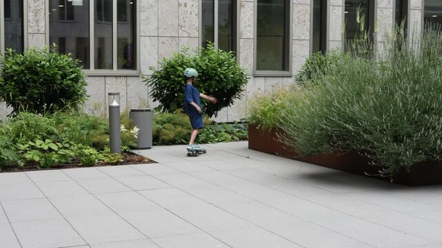 Young boy in helmet rides skateboard on paved urban area with greenery