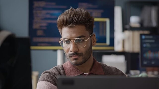 Rack focus shot of concentrated Indian young male developer wearing glasses working on computer in IT office, analyzing data on screens. Perfect for software development and tech support commercials