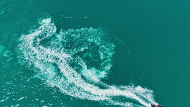 A person is on a jet ski creating waves in bright blue water on a sunny day close to the shoreline. The movement forms patterns in the water as the sun shines above.