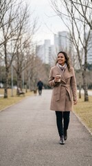 Fototapeta premium Mulher sorrindo enquanto caminha em parque urbano com árvores secas durante inverno, vestindo casaco elegante e segurando bebida quente