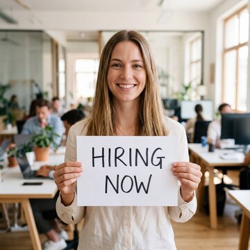 Smiling young caucasian woman holding a hiring now sign in a busy modern office
