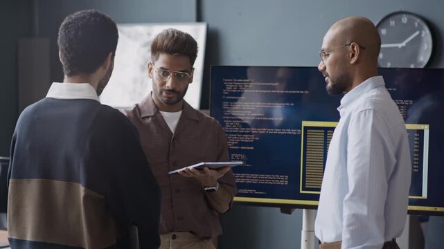Medium shot of young Indian male IT specialists discussing software code on monitor while brainstorming in office. Perfect for tech startup promotions and corporate training