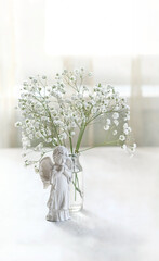 Obraz premium Beautiful angel baby figurine and bouquet of white gypsophila flowers in glass vase on table, abstract light background. symbol of faith, pray, believe, christianity. romantic, lovely atmosphere