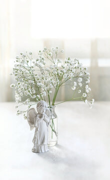 Beautiful angel baby figurine and bouquet of white gypsophila flowers in glass vase on table, abstract light background. symbol of faith, pray, believe, christianity. romantic, lovely atmosphere