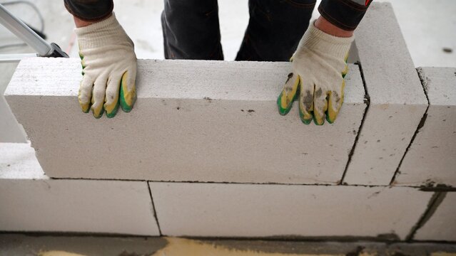 Construction worker in gloves placing a large aerated concrete block while building a wall at a construction site.