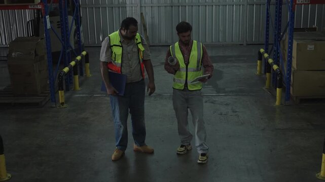 Two men conduct detailed warehouse inspection amidst stacked boxes and shelving units within darkened industrial facility, assessing safety barriers during an equipment maintenance process.
