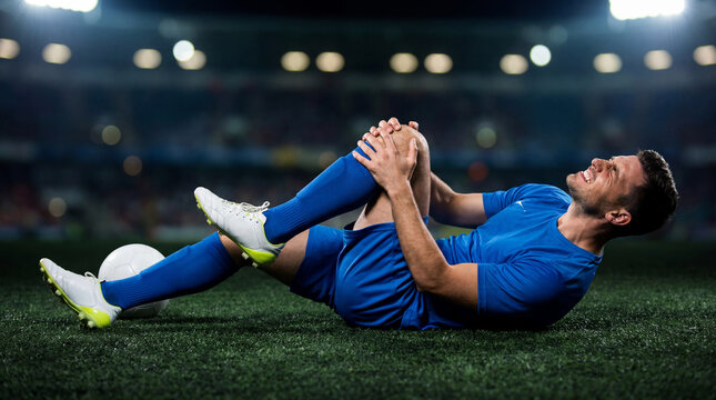 A soccer player lying on the stadium field clutching his injured knee in severe pain during a night match