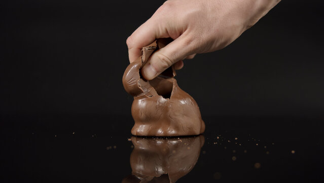Hand breaking dark chocolate egg against black background with glossy surface and small crumbs flying