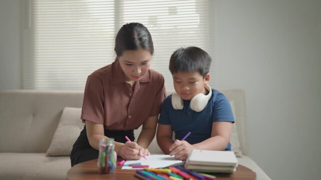 Happy asian mother helping her son with homework, drawing with colorful markers on paper while sitting on the sofa in the living room, showing a loving and supportive family relationship