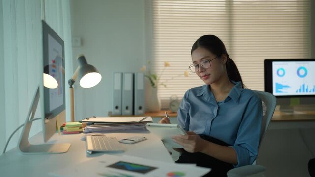 Thoughtful asian businesswoman sitting at her desk, brainstorming new project ideas and taking notes in her notebook while working overtime in a modern office during the evening hours
